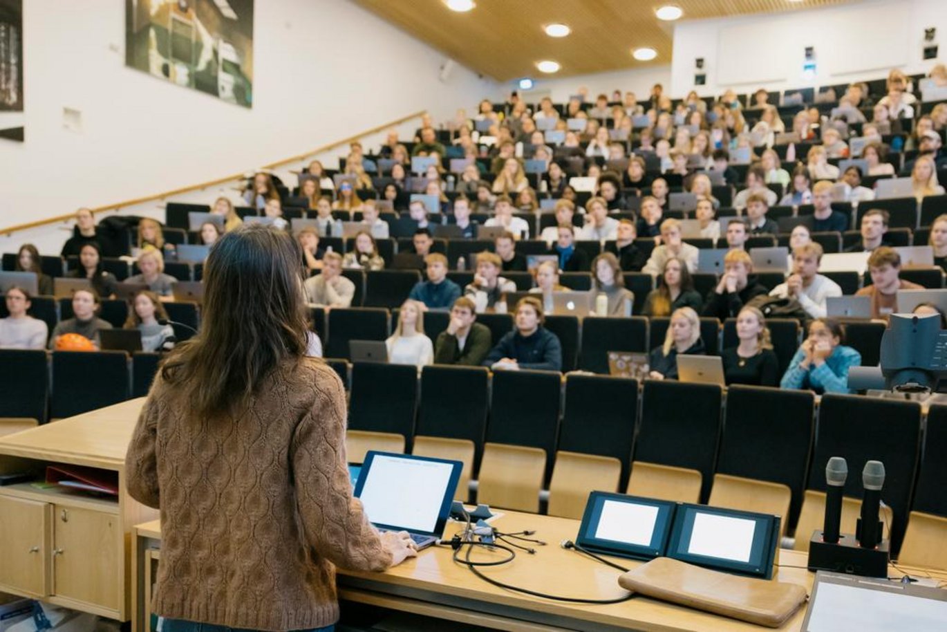 Foto af et fyldt auditorium, hvor en underviser står ved en talerstol med en præsentation foran mange kursusdeltagere på rækker af sæder.