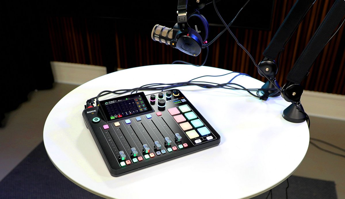 Close-up of a mixer with a display, faders, and coloured buttons on a round table, with a microphone and headphones visible in the background.
