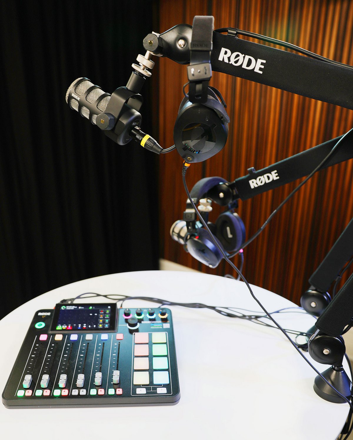 Podcast setup with two Rode Procaster microphones on boom arms, two pairs of headphones, and a mixer on a round table.