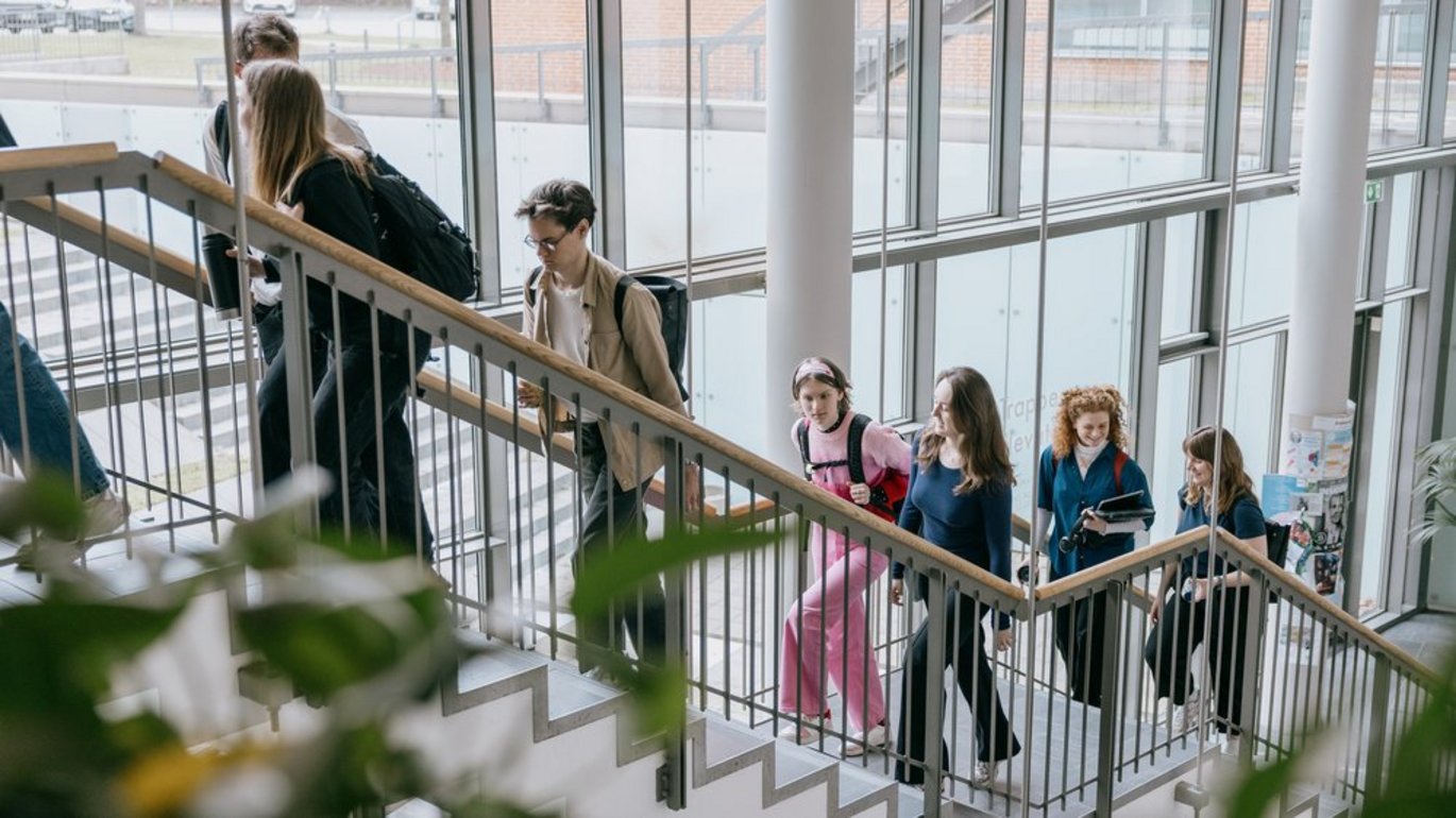 Studerende går op ad en trappe i en moderne universitetsbygning med store vinduespartier og masser af dagslys.