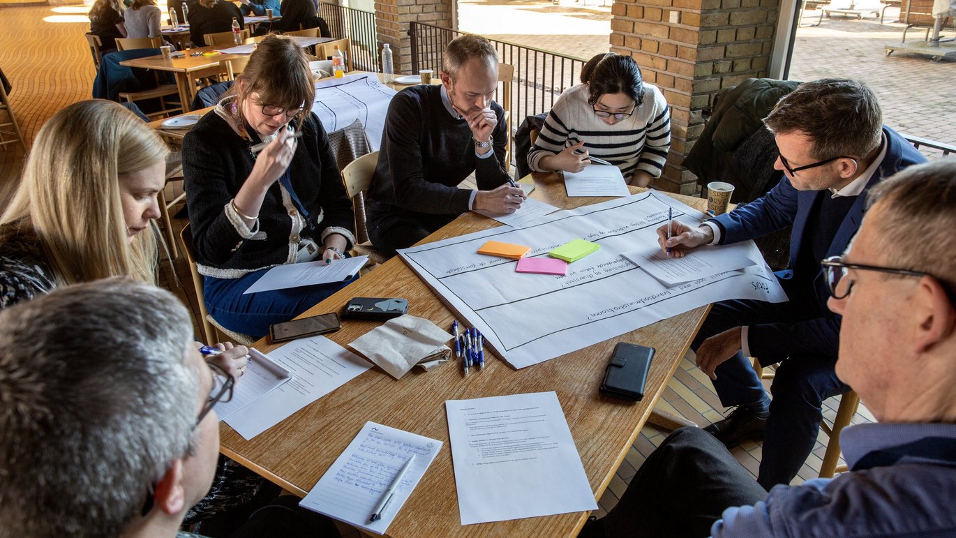 Photo of people sitting around a table, working together in groups with papers and notes.
